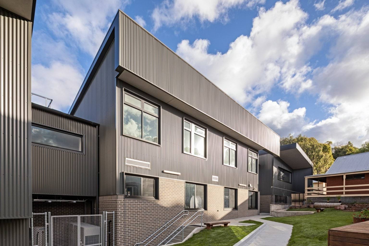 The exterior of a two-storey school building with grey corrugated cladding and brickwork at the base. Large windows overlook landscaped paths and grassed areas.