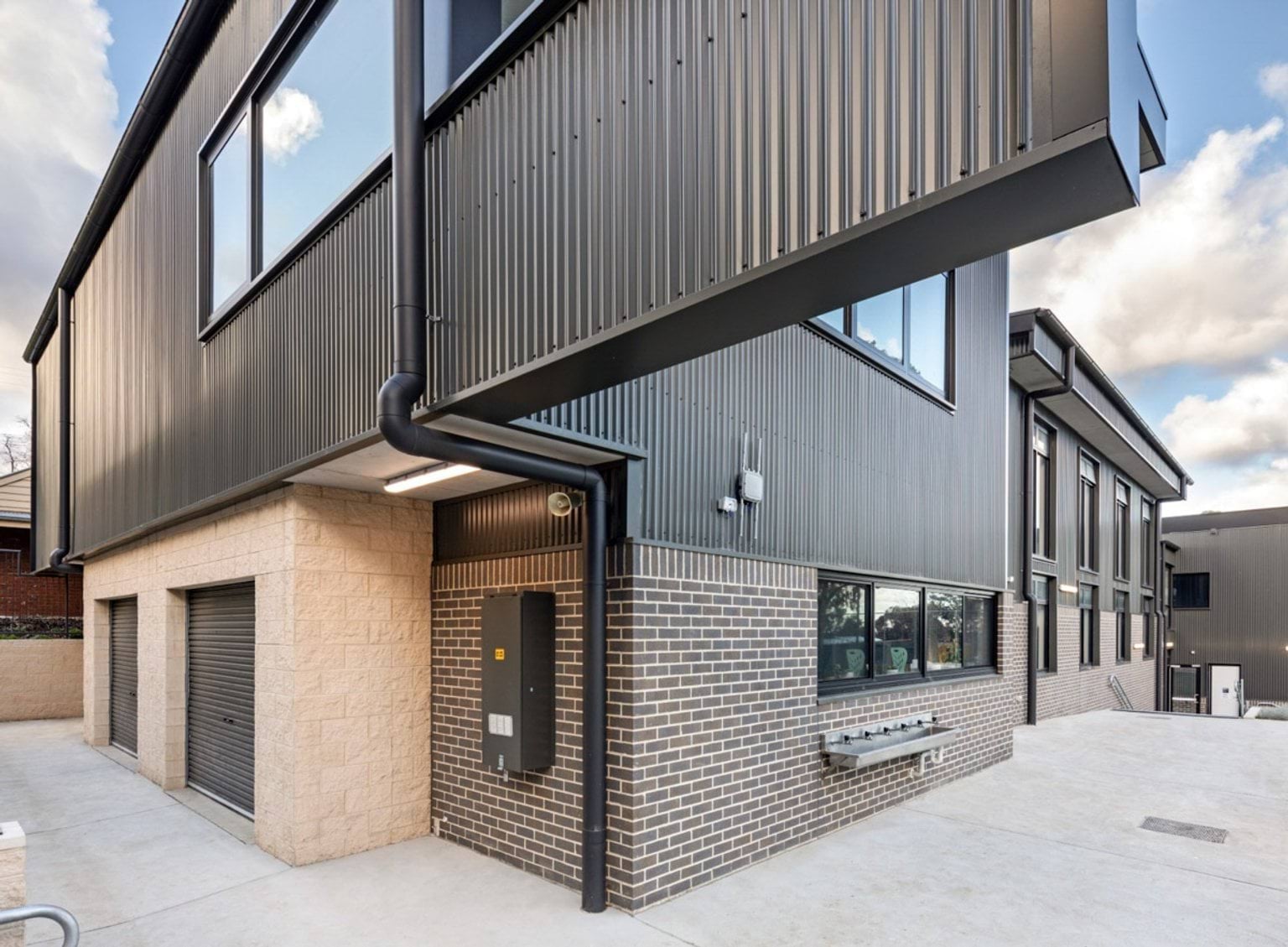 Corner view of a school building with grey corrugated cladding and brickwork. A concrete courtyard and outdoor sinks are visible along the wall.