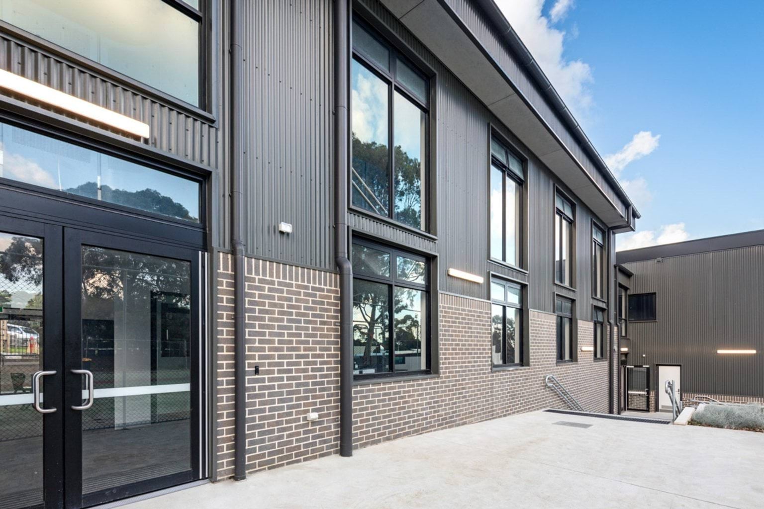 Close-up of a school building with grey corrugated cladding, brickwork, and large windows. A concrete path and steps lead to the entrance.