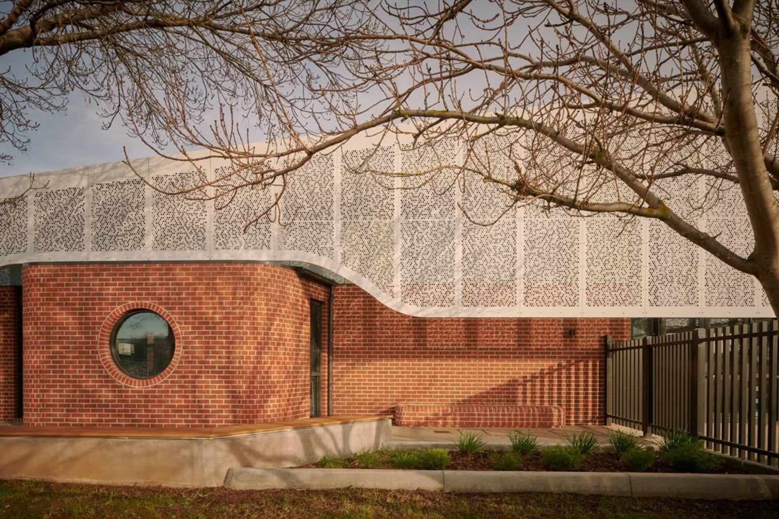 Close-up of a school building’s curved white patterned facade over red brick walls, with a round window and landscaped garden beds in front.