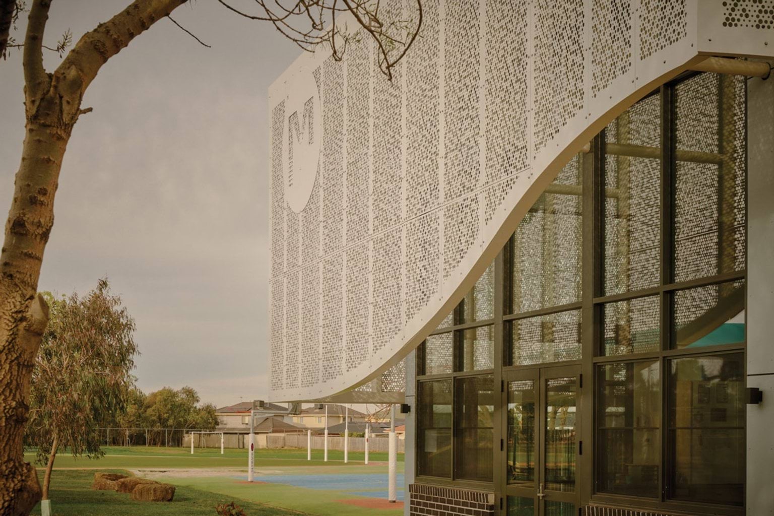Detail of a school building’s curved white patterned facade above large glass windows, with views of the sports field in the background.