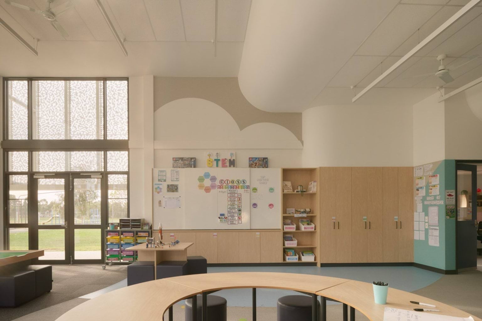 A bright STEM classroom with curved timber tables, round stools, and large windows with patterned screens. A whiteboard and timber storage cabinets line the back wall.