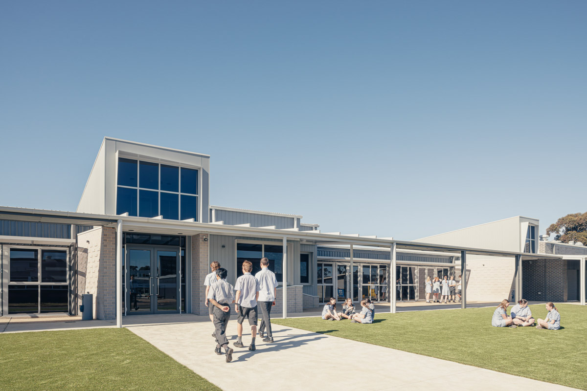 Modern school building with large windows and covered walkway, students walking and sitting on grassed courtyard