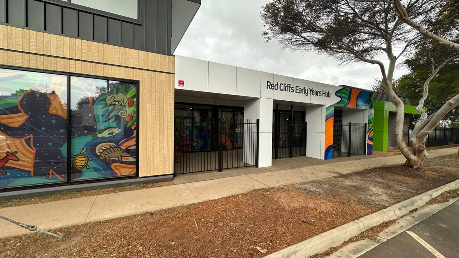 The exterior of the early years hub with a white facade and bold Aboriginal-inspired artwork panels. Large windows display colourful designs of animals and nature, and a black metal fence runs along the entrance.