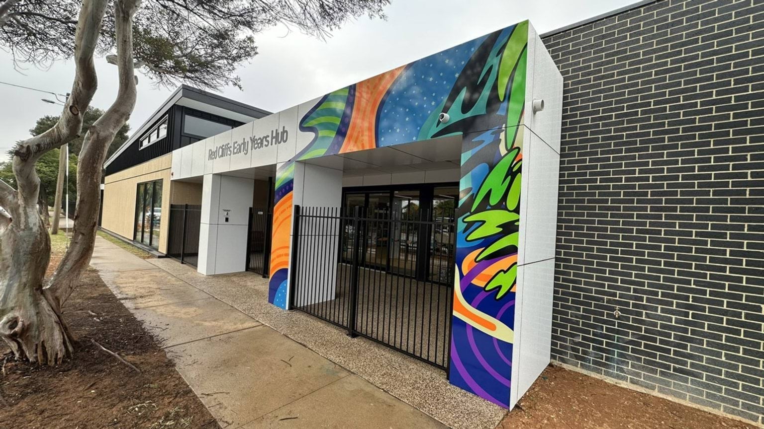 The entrance of the early years hub with a white facade and colourful art panels. A black metal gate secures the entryway.