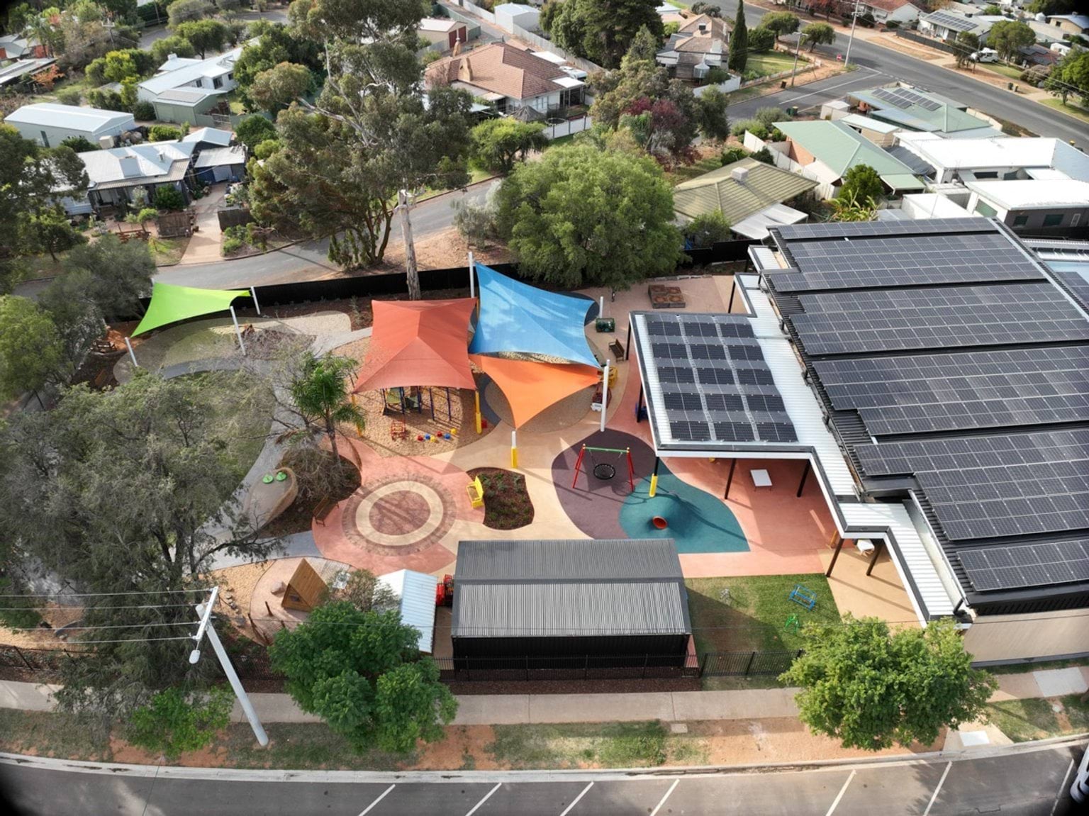 An aerial view of the outdoor play area with colourful shade sails, circular paving patterns, and landscaped garden beds. The building roof is covered with solar panels.