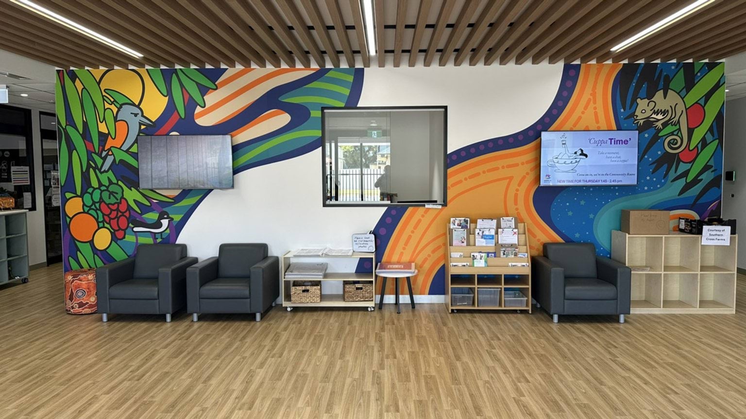 A reception area with grey armchairs, a timber-look floor, and a wall featuring vibrant artwork. A TV screen and brochure stand are mounted on the wall.