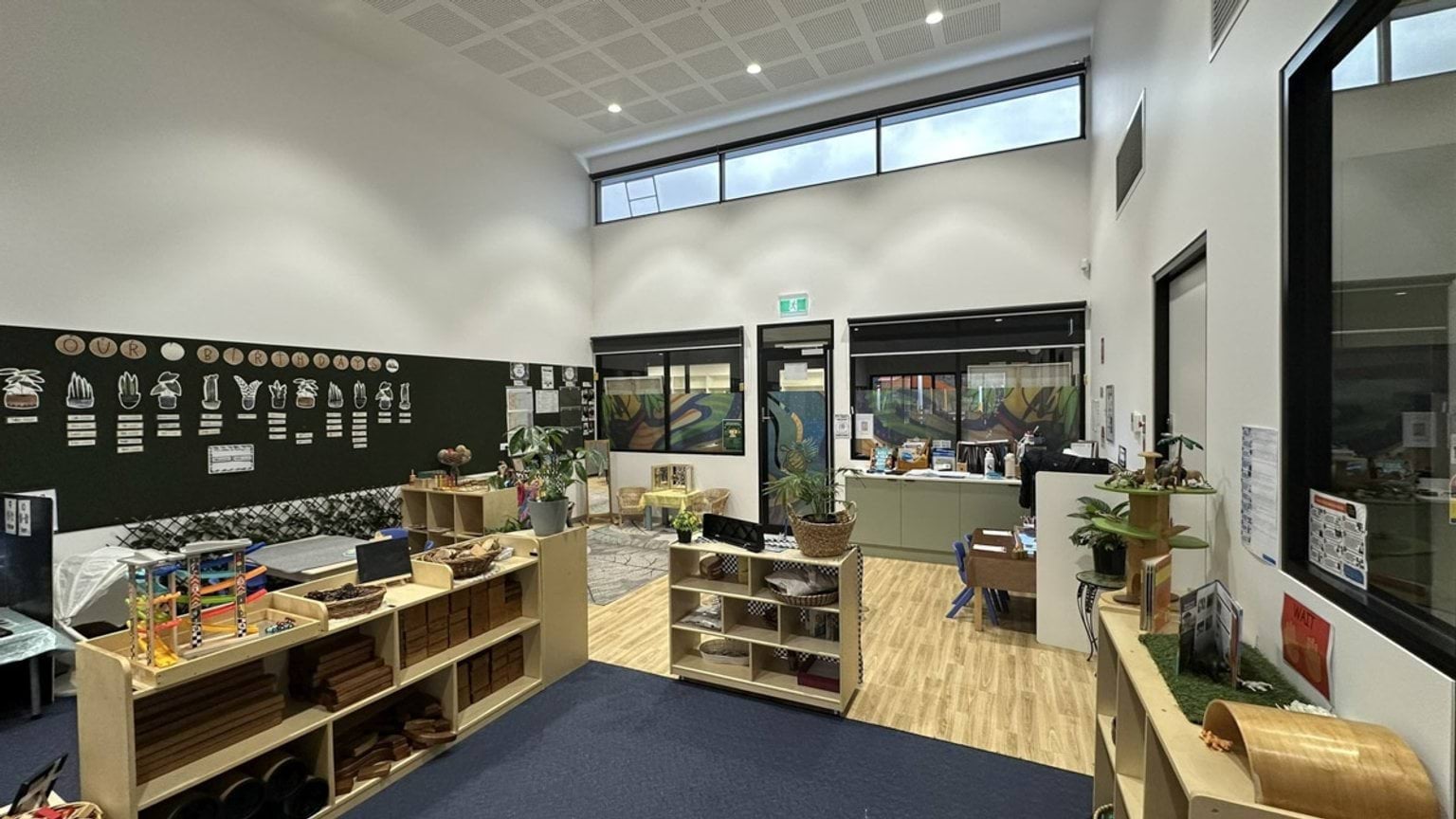 A kindergarten classroom with timber-look flooring, low shelving, and plants. The walls display educational posters, and large windows bring in natural light.