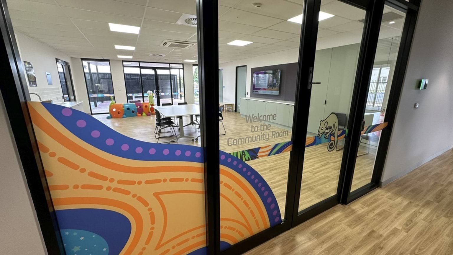 A bright community room with timber-look flooring, tables and chairs, and large glass doors featuring colourful Aboriginal-inspired designs. Soft play blocks and toys are visible near the windows.