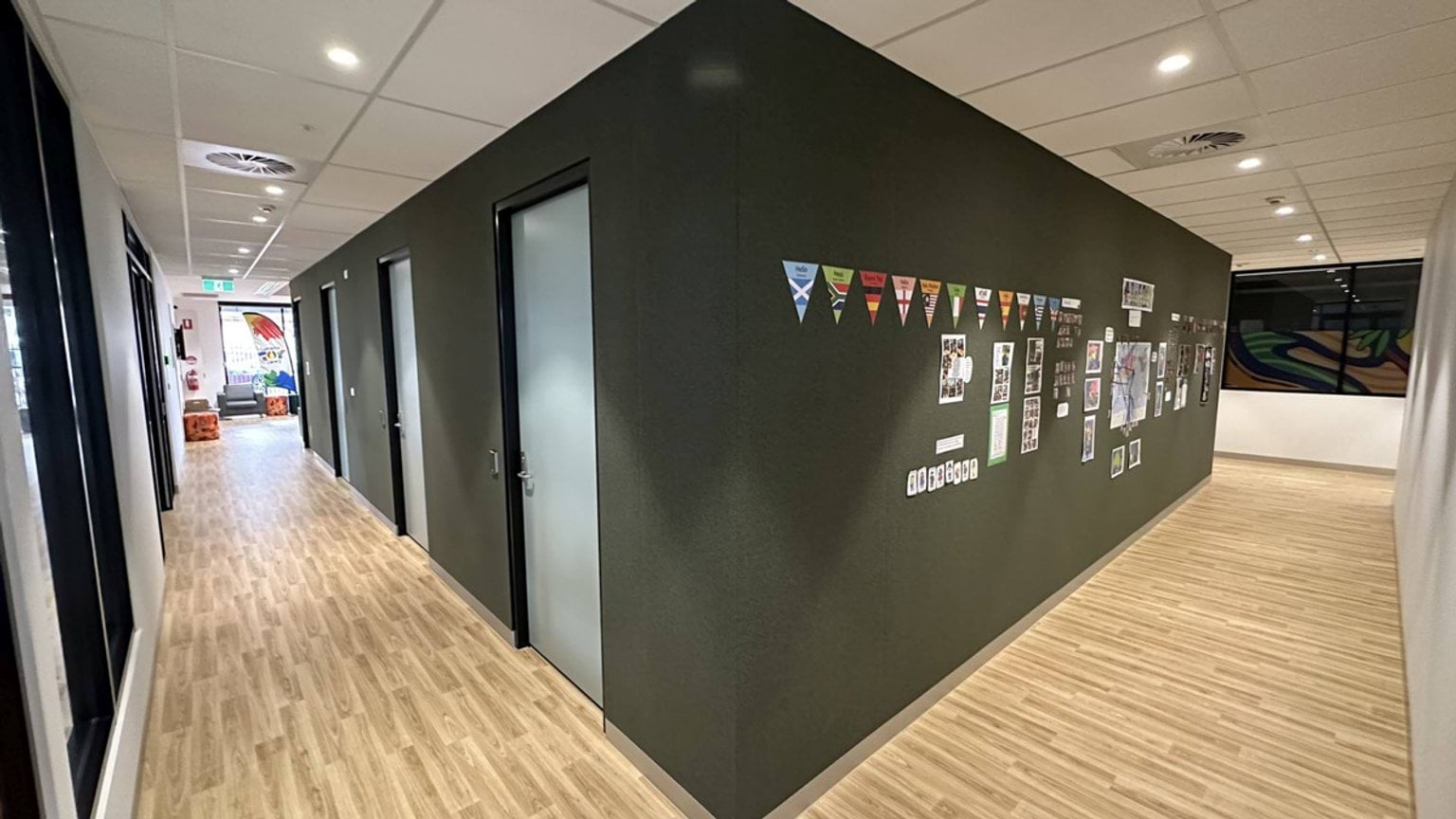 A hallway with timber-look flooring and a dark green wall displaying flags and photos. Several doors lead to adjoining rooms, and natural light enters from the far end.