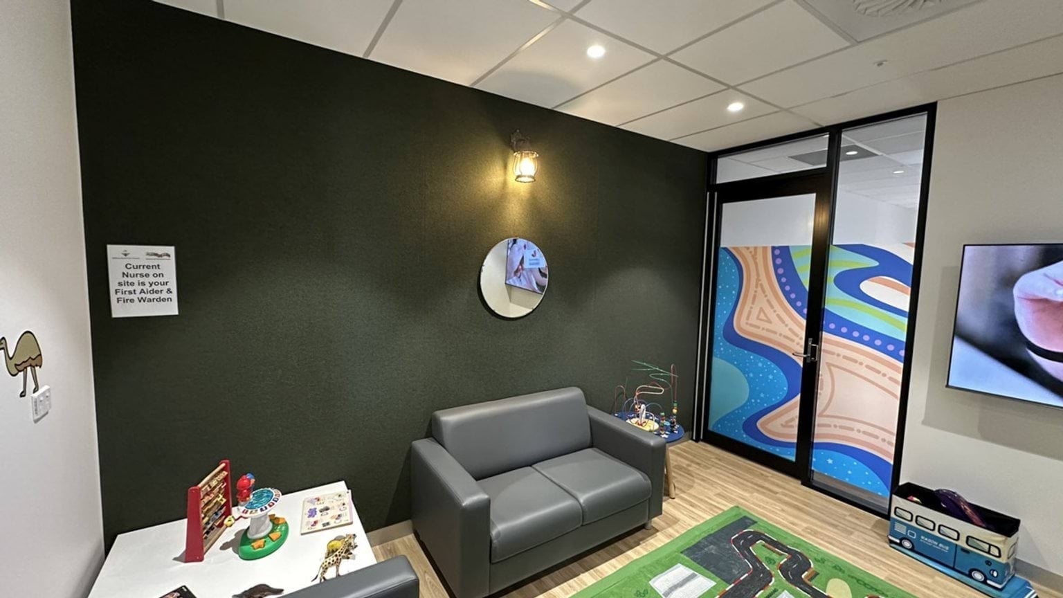 A waiting area with a dark green feature wall, a grey couch, and a play mat with a road design. Toys are arranged on a small table, and the glass door has colourful artwork.