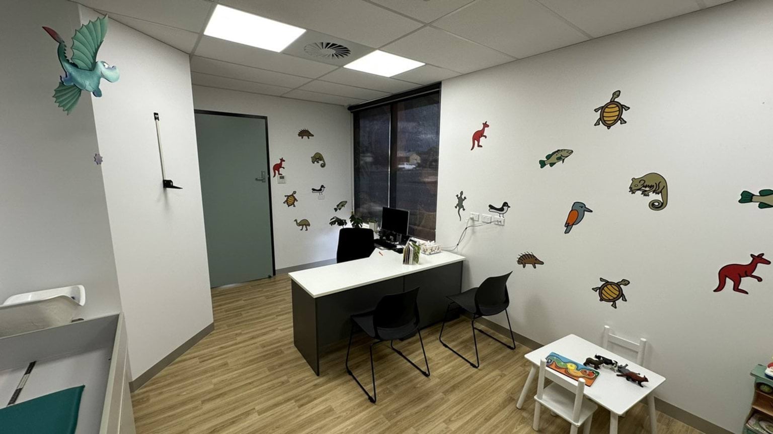 A consultation room with a desk, two chairs, and a small play table. The walls are decorated with colourful animal decals, and the floor has a timber-look finish.
