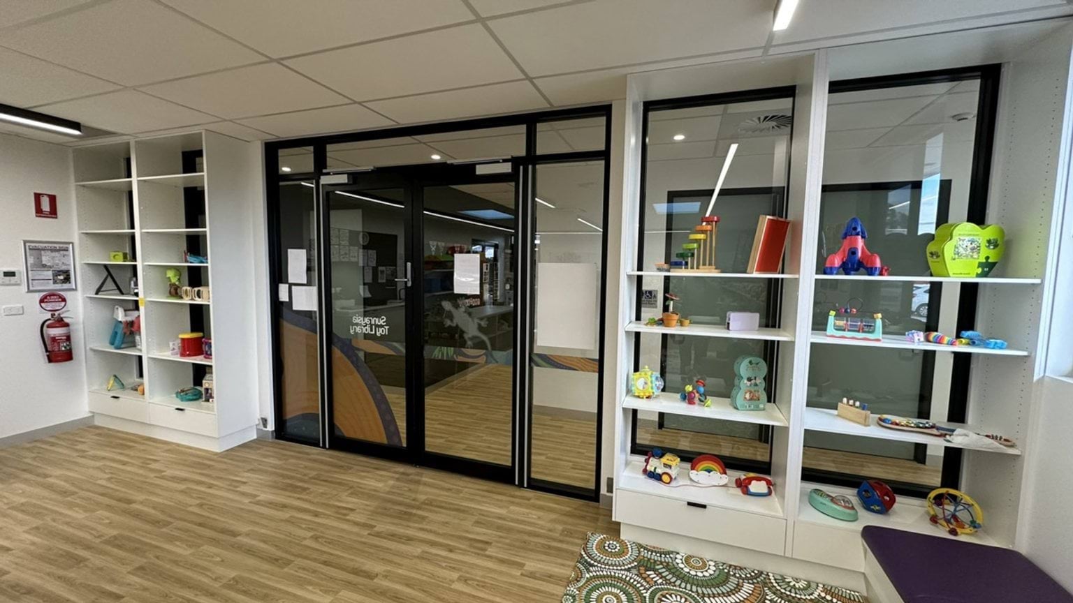 A toy library area with white shelving filled with colourful toys and games. Glass doors feature artwork, and the floor has a timber-look finish.