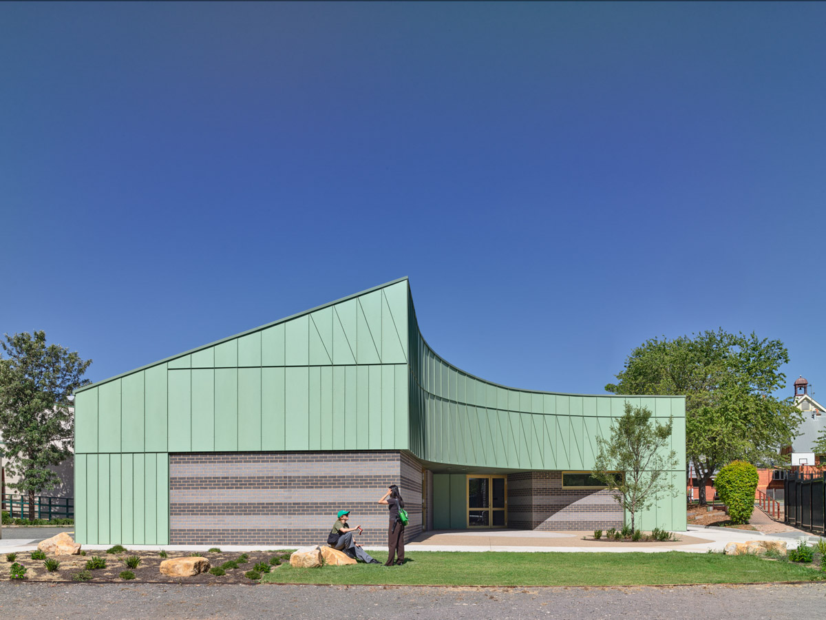 A modern school building with green cladding and grey brickwork, featuring a curved roofline and landscaped garden beds under a clear blue sky.