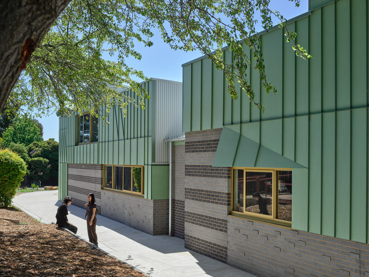 The exterior of a modern school building with green metal cladding, grey brickwork, and yellow-framed windows. A concrete path runs alongside landscaped garden beds and trees.