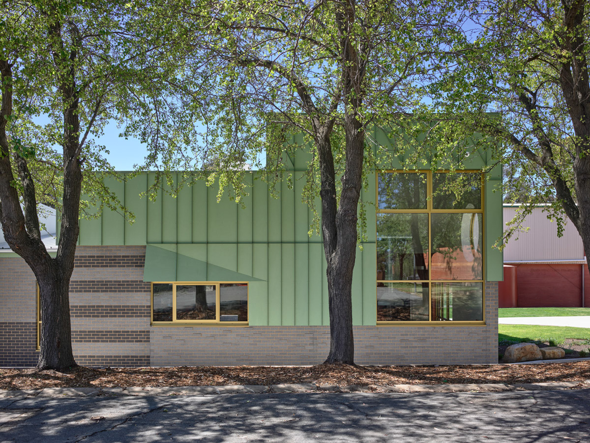 Side view of a modern school building with green cladding, grey brickwork, and yellow-framed windows, partially shaded by mature trees.