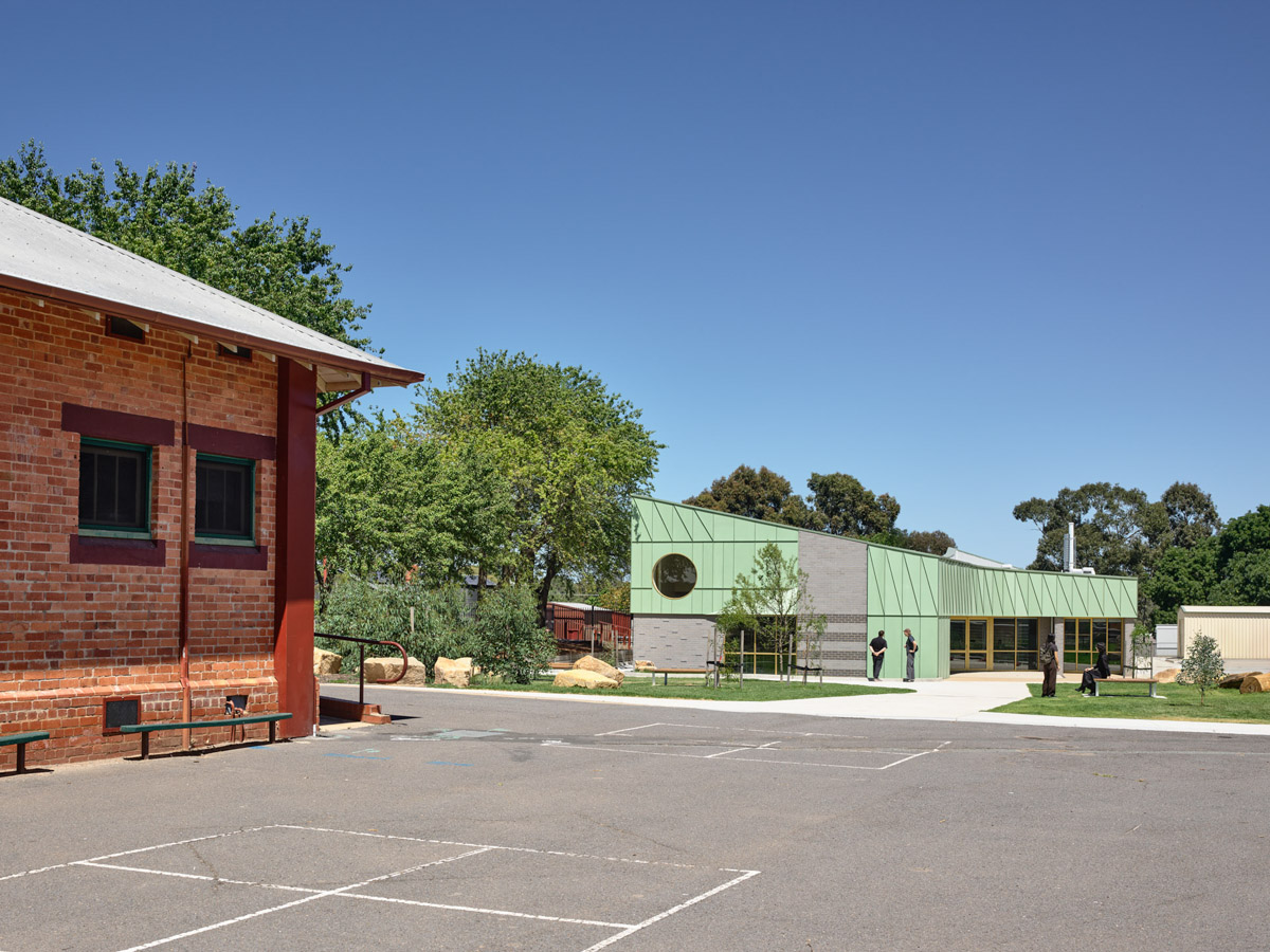 A school courtyard with a heritage red brick building on one side and a modern green-clad building on the other. Trees and landscaped garden beds frame the space.