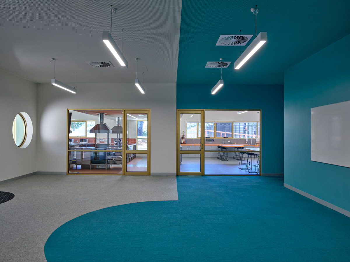 An open learning space with teal and grey carpet, white walls, and suspended linear lights. Glass doors lead to adjoining rooms with high benches and stools.