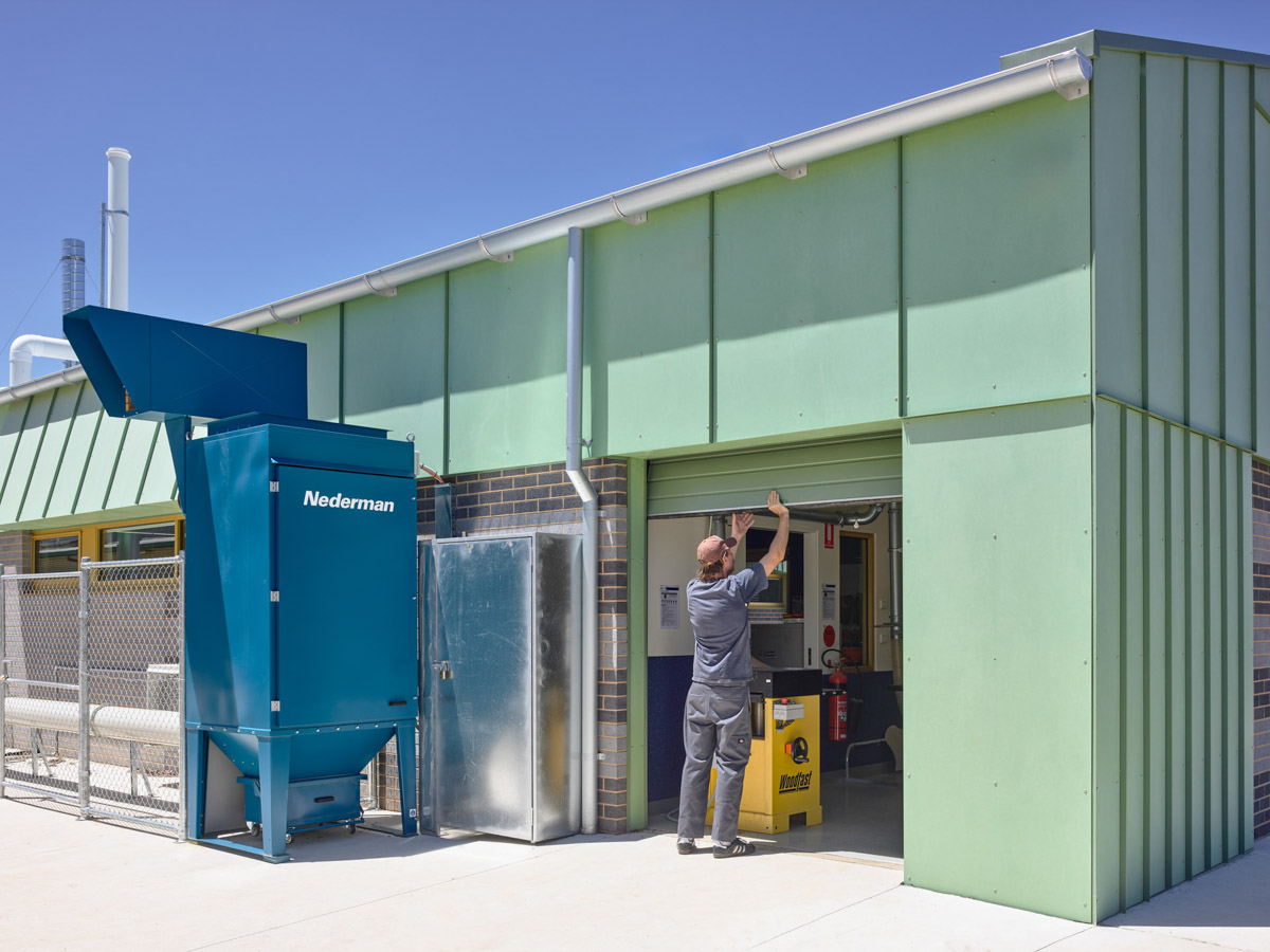 A school workshop building with green cladding and grey brickwork. A large blue dust extraction unit is installed outside near a roller door.
