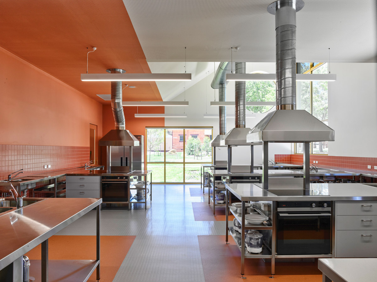 A kitchen classroom with stainless steel benches, ovens, and large range hoods. Orange walls and tiled splashbacks contrast with the bright interior.