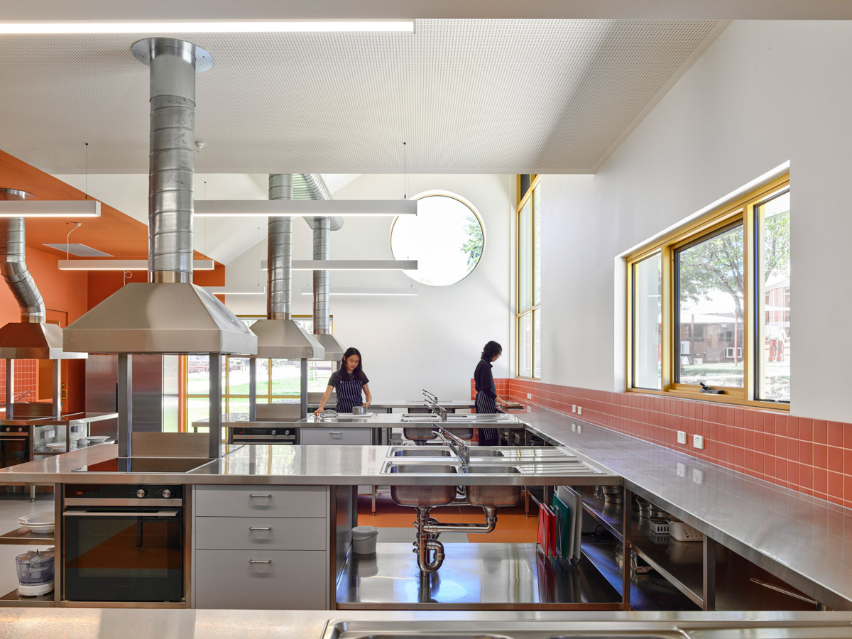 A commercial kitchen classroom with stainless steel benches, ovens, and large range hoods. Orange feature walls and round windows add colour to the space.