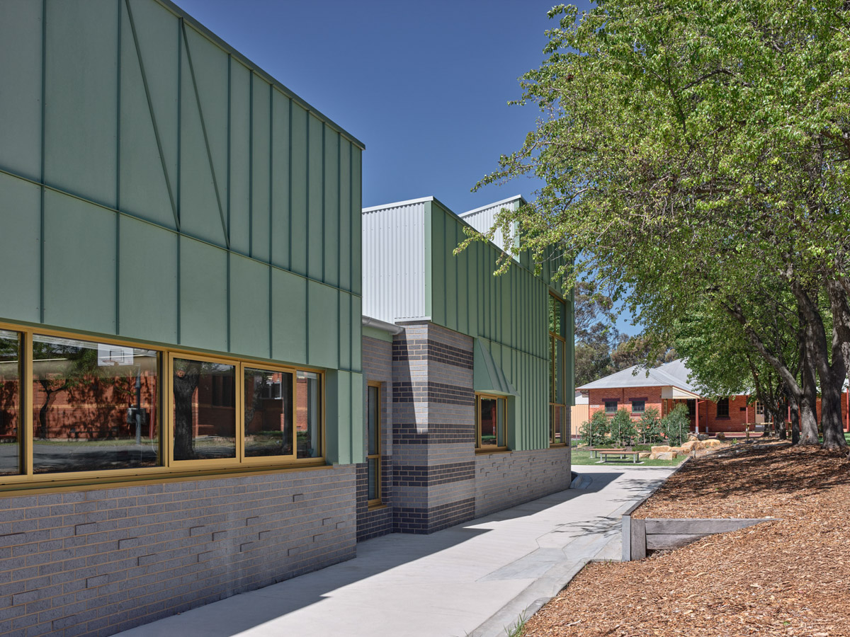 The exterior of a modern school building with green cladding and grey brickwork, framed by mature trees and landscaped garden beds.