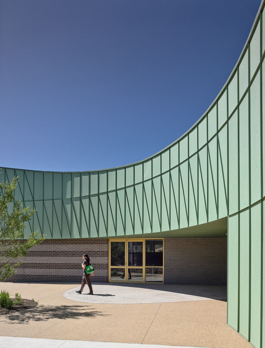 A curved entrance to a school building with green cladding, grey brickwork, and yellow-framed glass doors. Landscaping surrounds the paved entry area.