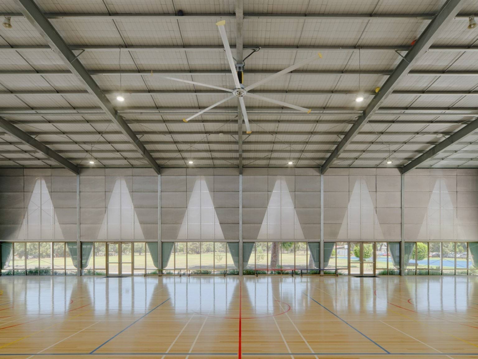 An indoor sports court with polished timber flooring, marked for multiple games. The walls have triangular panels and full-height windows, and a large ceiling fan hangs from the roof trusses.