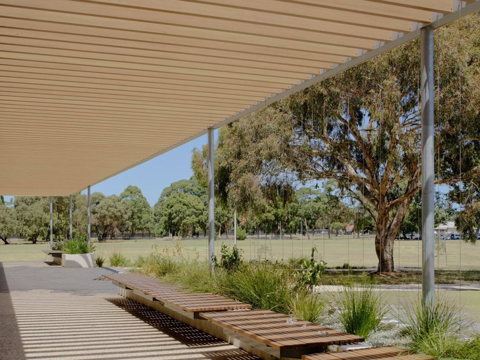 A covered outdoor seating area with timber benches and landscaped garden beds, overlooking a grassed sports field and mature trees.