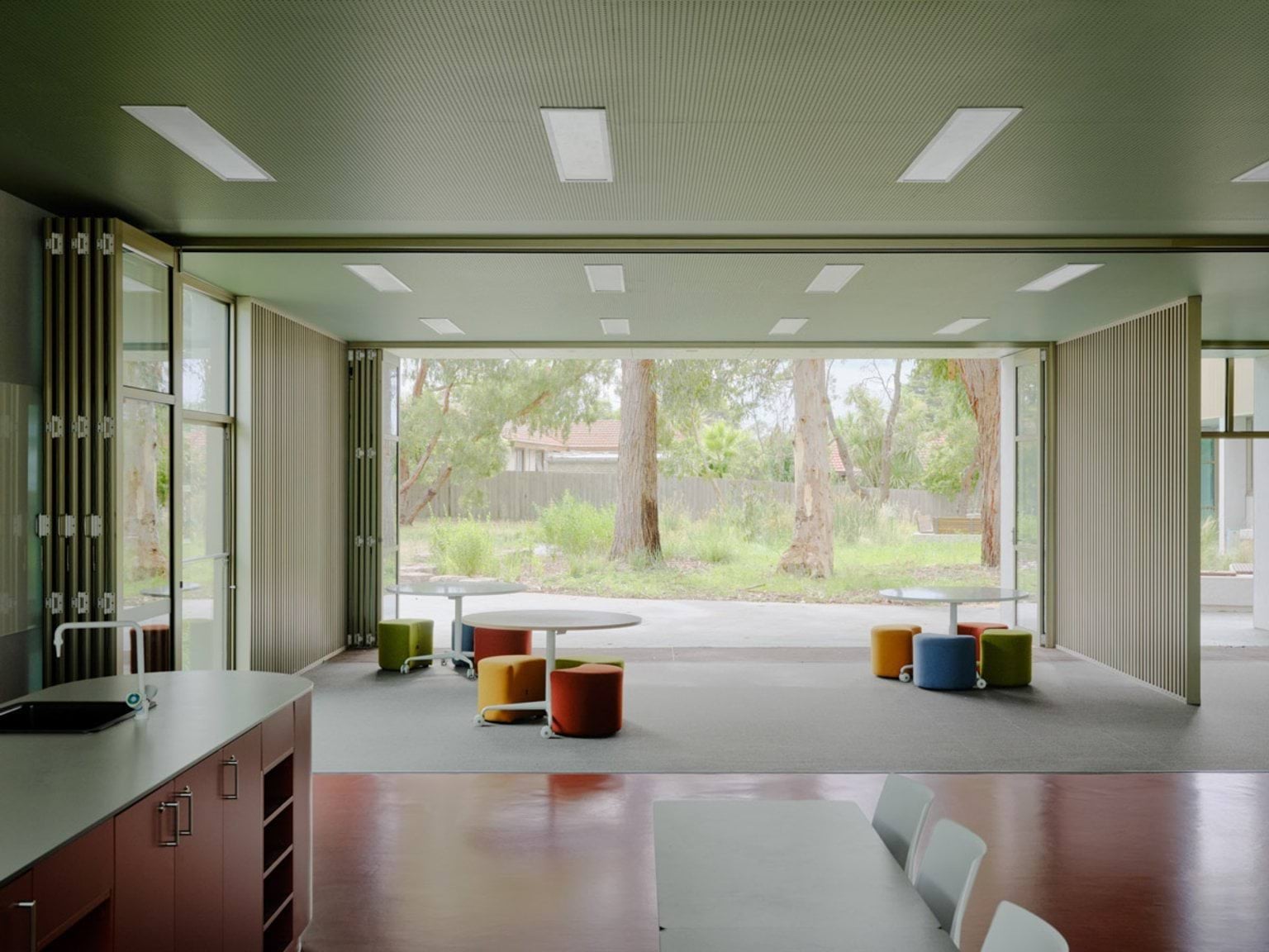 A flexible learning space with folding doors opening to an outdoor area. Round tables and colourful stools are arranged on grey carpet, with large windows providing natural light.