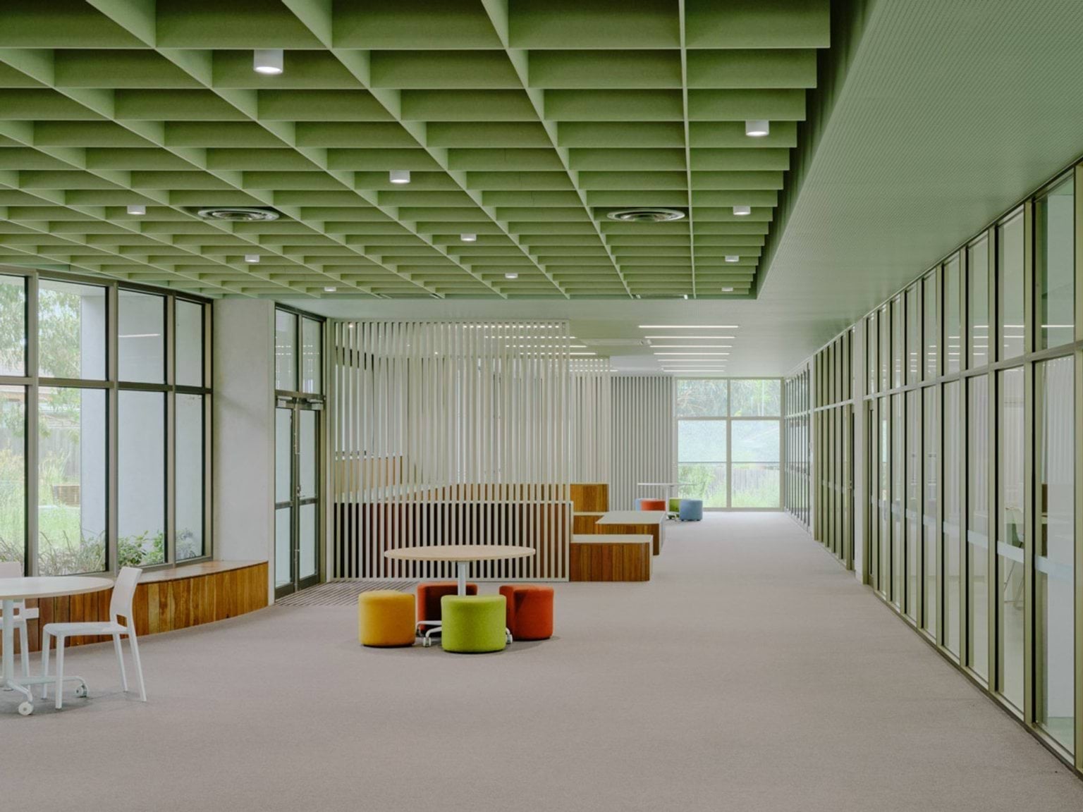An open learning space with a green grid ceiling, glass walls, and soft seating in bright colours. Timber benches and tables are arranged throughout the area.