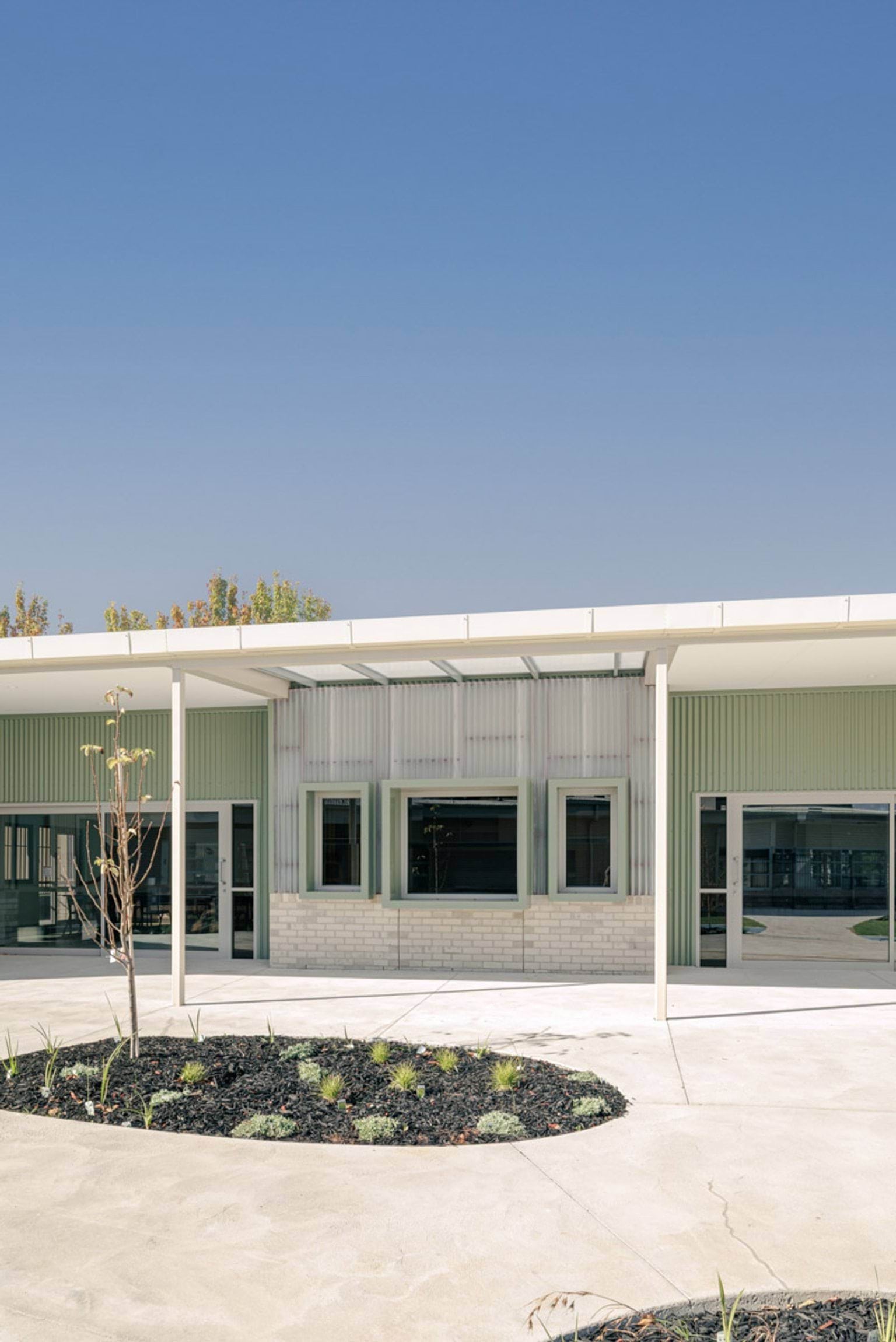 The exterior of a school building with green and cream cladding, light brickwork, and a covered walkway. A circular garden bed with young plants is in the foreground.