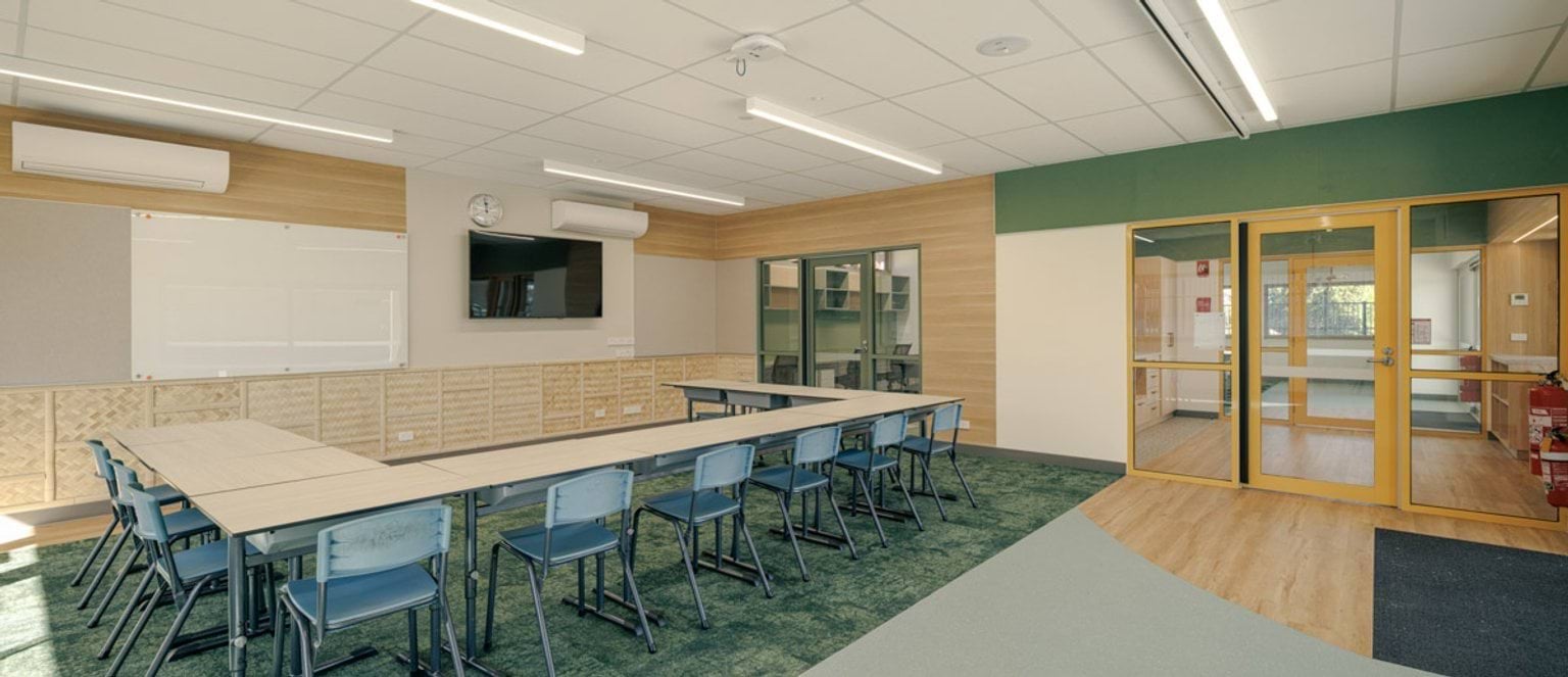 A classroom with U-shaped tables and blue chairs on green carpet. The walls have timber panels, a whiteboard, and a mounted screen. Double glass doors lead to another space.