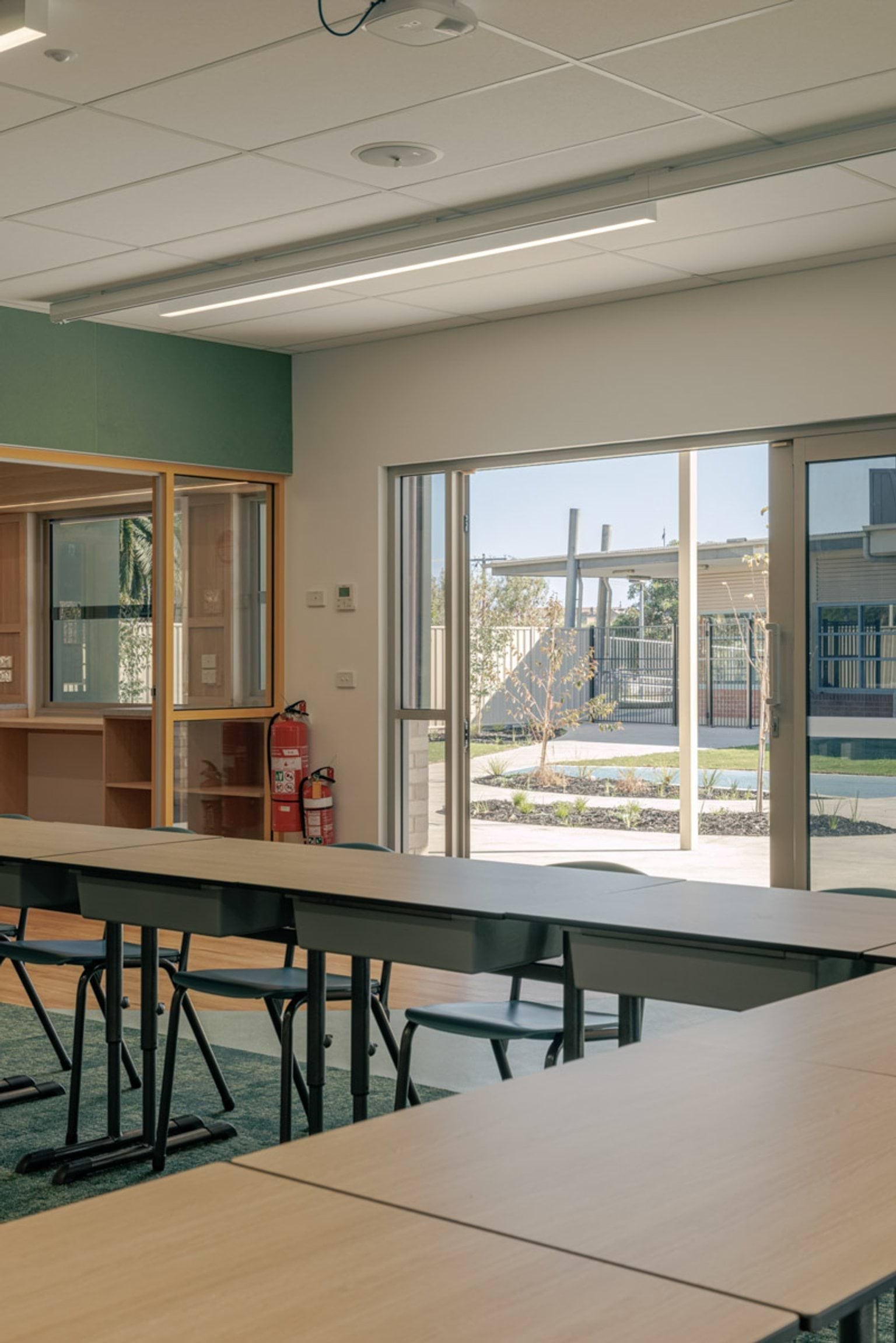 Interior view of a classroom with tables and chairs, looking out through large glass doors to a landscaped courtyard with garden beds.