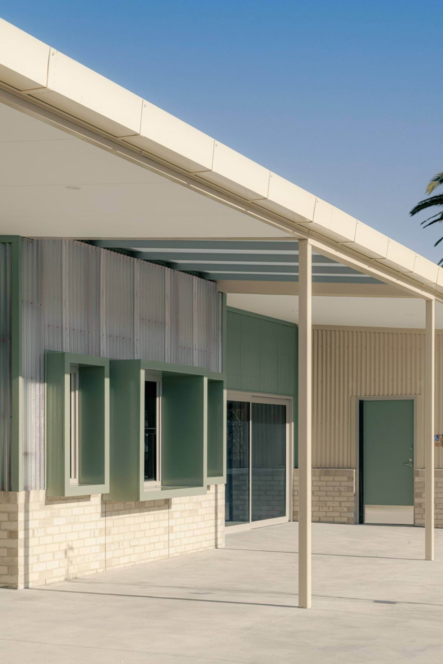 School exterior with a wide covered walkway, green box-shaped recesses, and corrugated cladding above light brick walls.