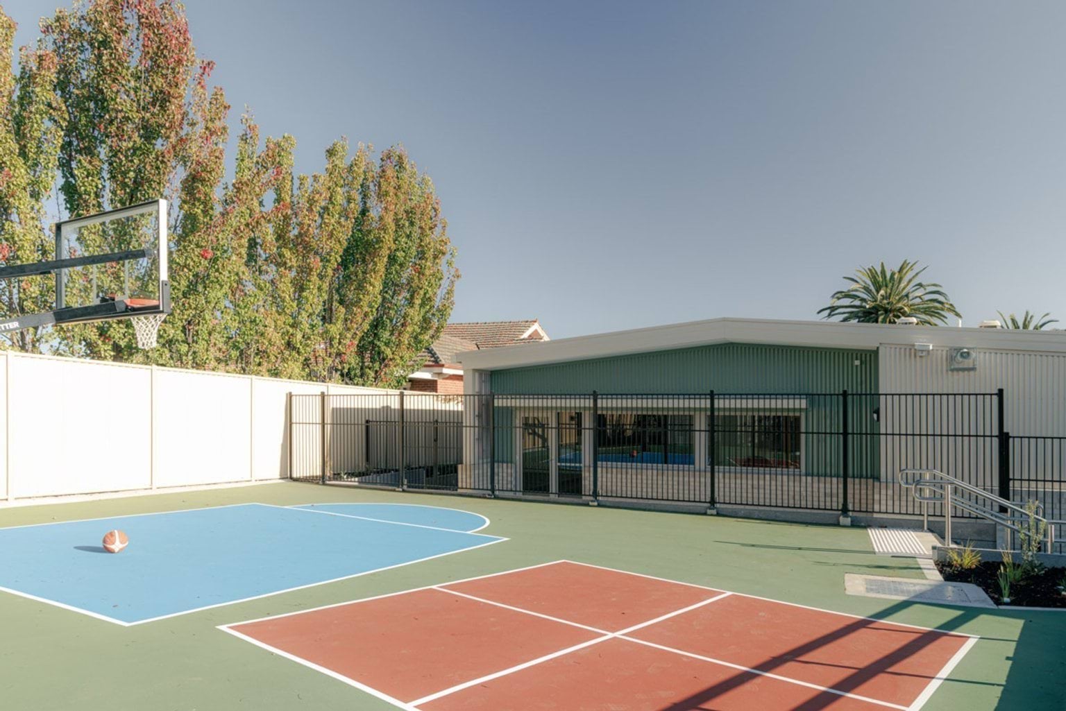 An outdoor sports court with blue and red playing surfaces and a basketball hoop, enclosed by a black fence. A school building and tall trees are in the background.