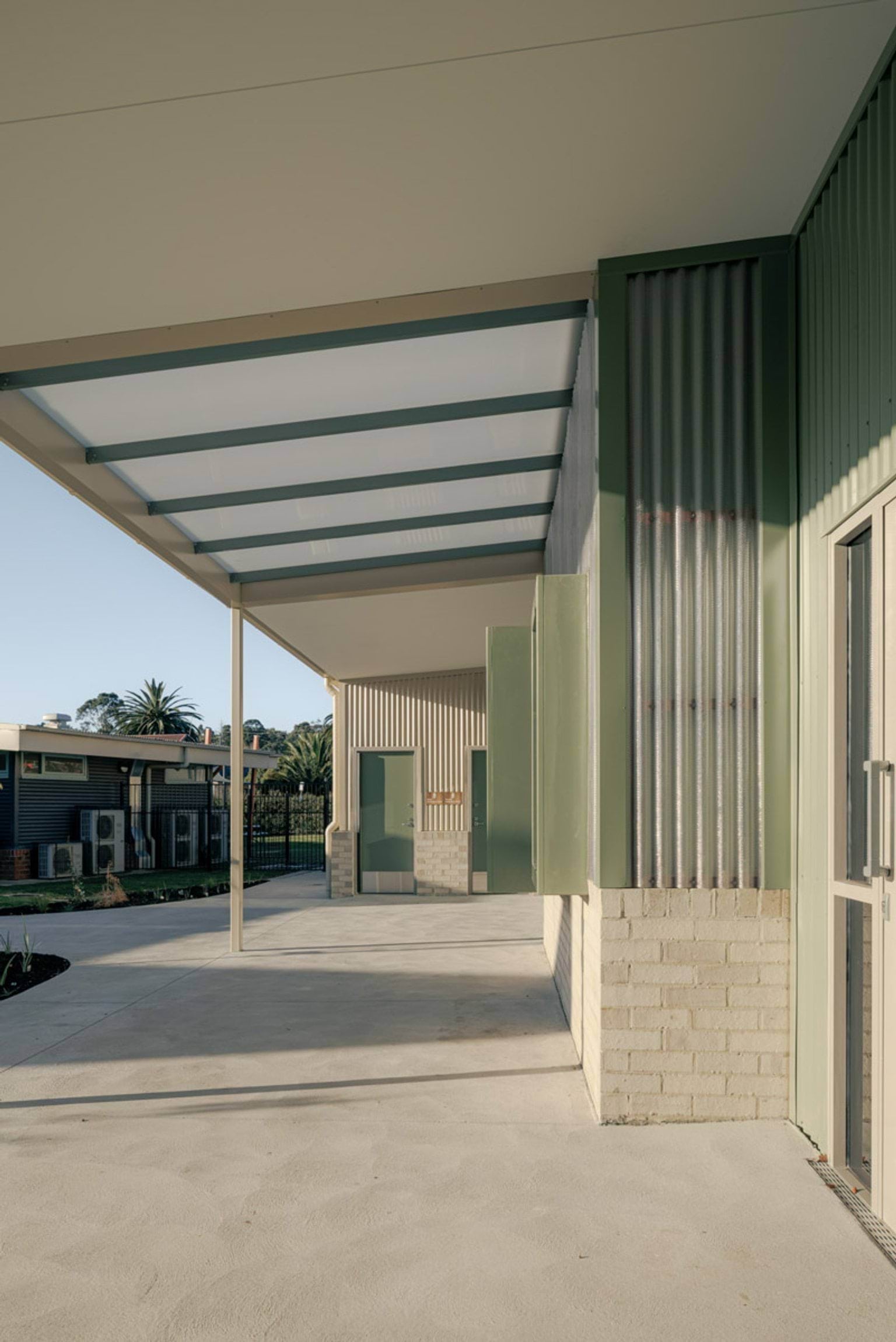A long covered walkway along the school building with green feature boxes and corrugated cladding. The floor is smooth concrete.