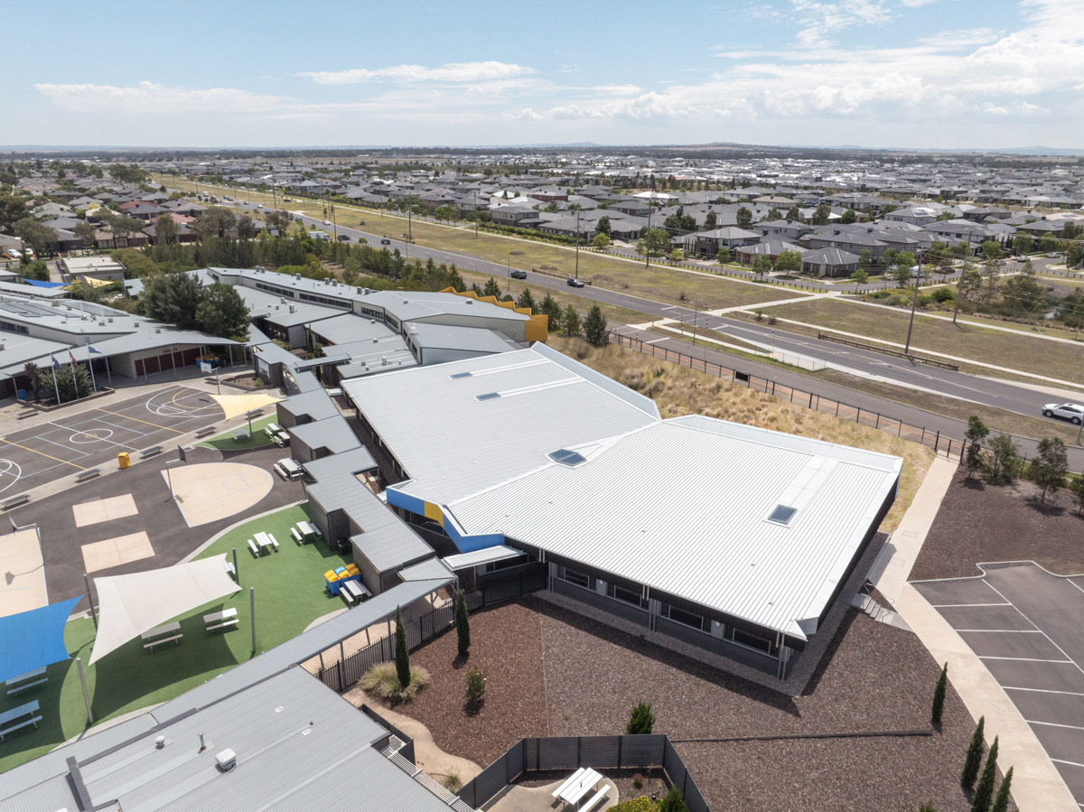 Aerial view of a modern school campus with multiple low-rise buildings, landscaped courtyards, and sports courts, surrounded by residential streets and open green spaces.