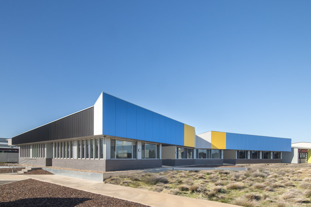 The exterior of a modern school building with bold blue and yellow cladding panels above grey brickwork, set against a clear blue sky.