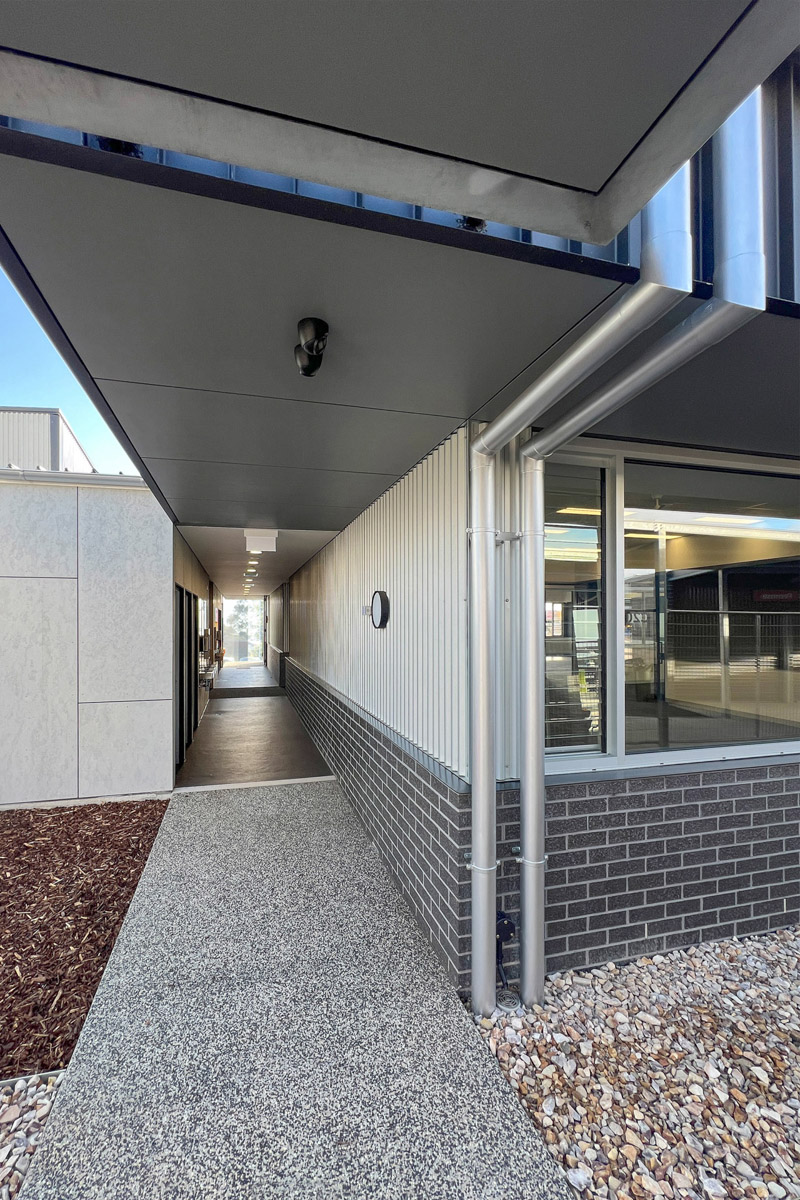A covered walkway with textured concrete flooring and steel posts, running alongside a school building with grey brick walls and large windows.