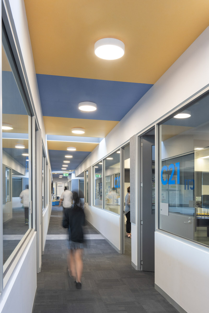 A hallway with a yellow ceiling panel and round lights, leading to a glass door at the end. Classrooms with large windows line both sides.