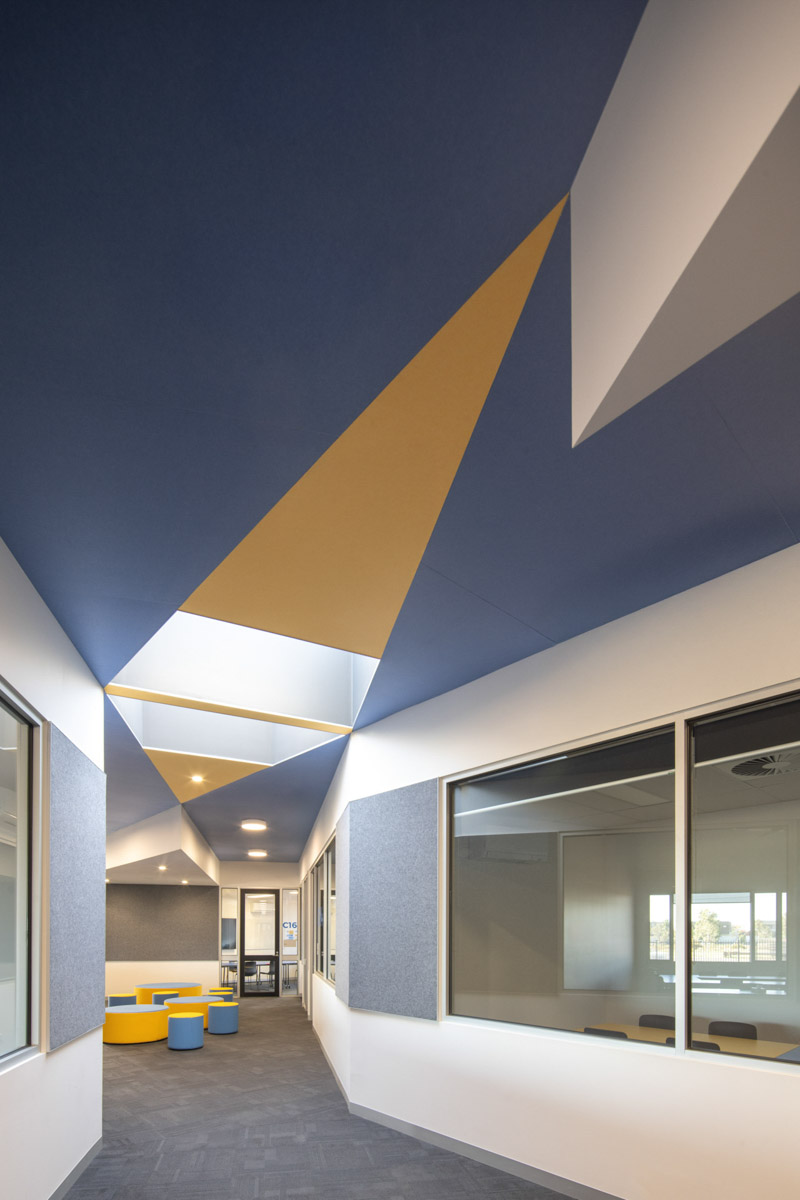 A hallway with a striking geometric ceiling design in blue and yellow, skylights, and glass walls opening to classrooms and breakout spaces with colourful seating.
