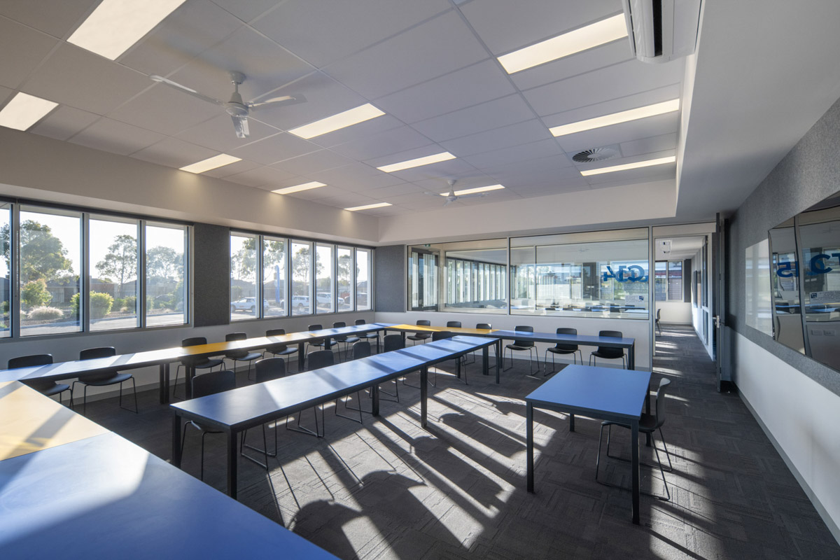 A classroom with blue and yellow tables arranged in a U-shape, black chairs, and large windows overlooking landscaped outdoor areas.