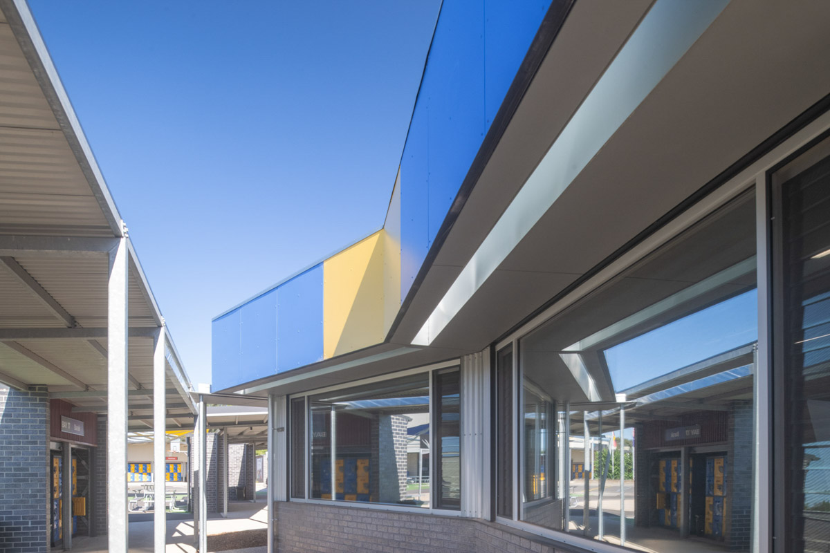 Exterior view of a school building with blue and yellow cladding panels above large windows, seen from a covered walkway with steel posts.
