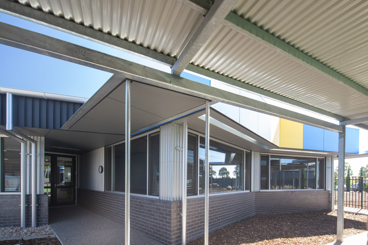 A covered outdoor walkway with corrugated metal roofing and steel posts, leading to a school building with grey brick walls and large windows.