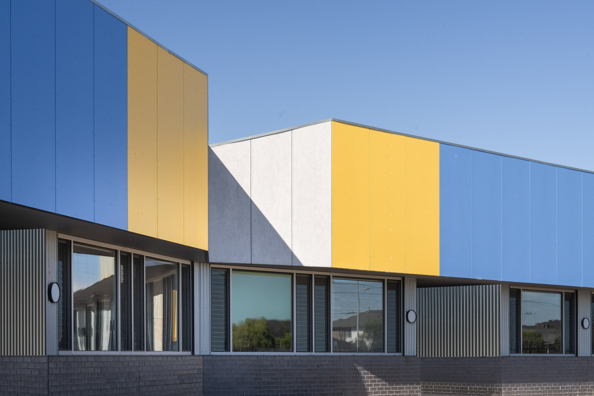 Detail view of a school building facade with alternating blue and yellow cladding panels above large windows framed by grey brickwork.