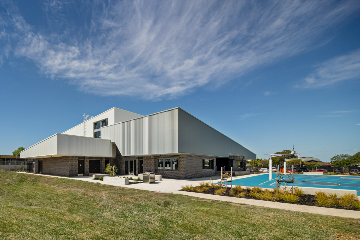 The exterior of a modern school building with grey corrugated cladding and dark brickwork, next to a blue outdoor sports court and landscaped garden beds.