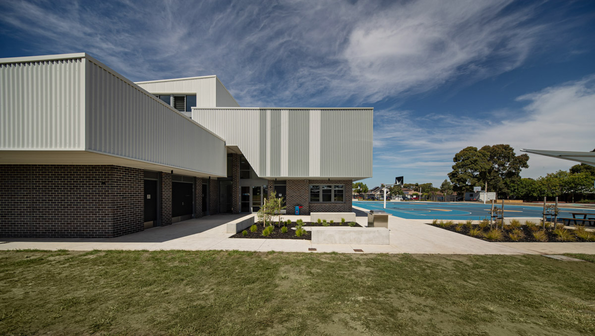 The exterior of a contemporary school building with grey corrugated cladding and dark brickwork. A landscaped entry with garden beds leads to the main doors, and a blue outdoor sports court is visible in the background.