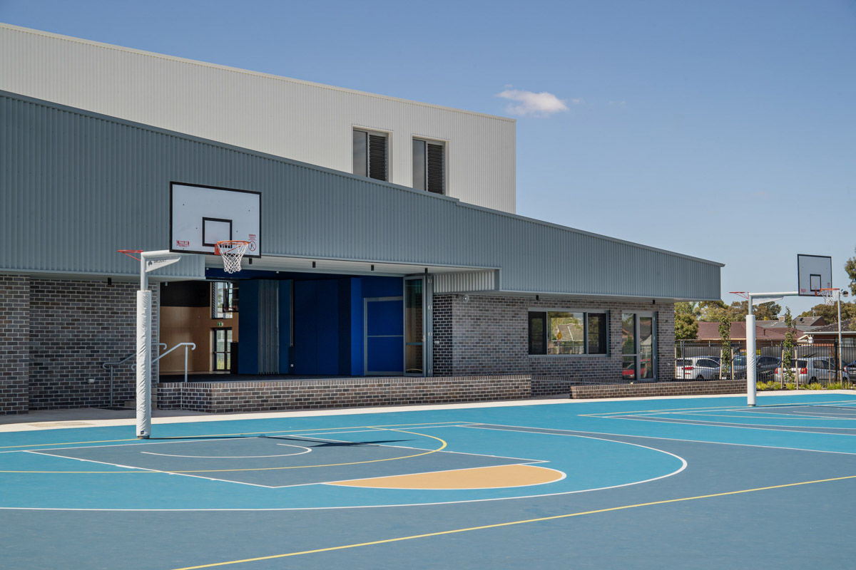 A blue outdoor basketball court with hoops in front of a modern school building featuring grey cladding and dark brickwork.