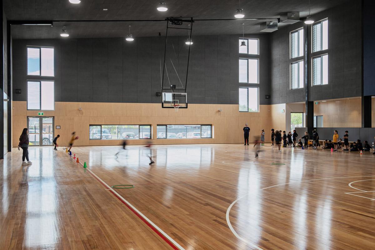 Students participating in physical activities on a polished timber indoor sports court with cones and hoops set up for games. Large windows provide natural light.